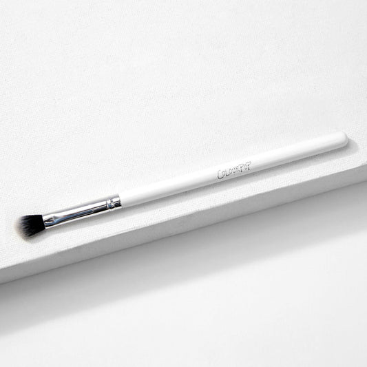 ColourPop Blending Brush makeup brush resting diagonally on a white textured surface, featuring fluffy angled black-to-white bristles, a silver ferrule, and a slim white handle, made for seamless eyeshadow blending.