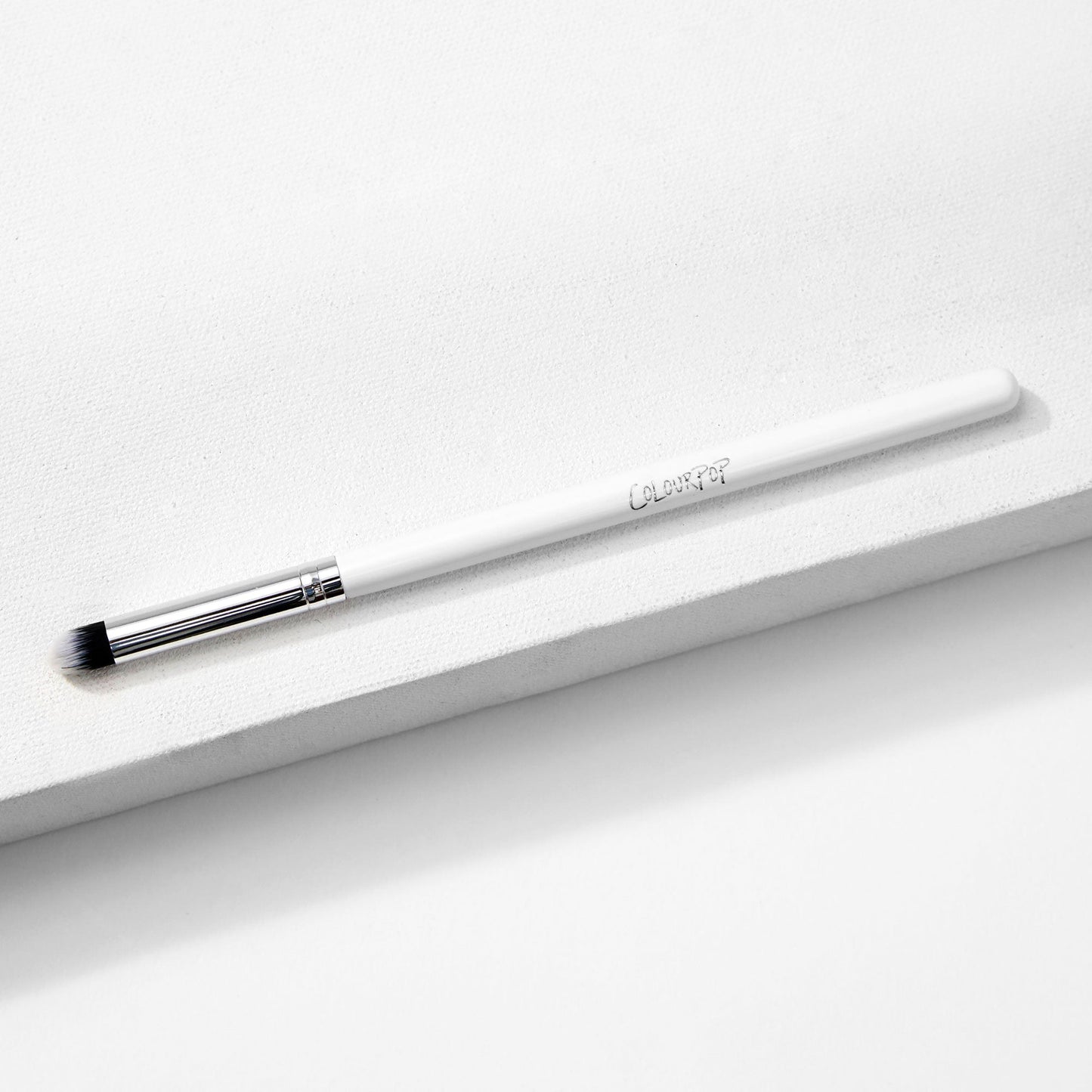 ColourPop Pencil Brush resting diagonally on a white textured block, a makeup brush with a slim white handle and logo, silver ferrule, and small tapered black-and-white bristles for precise eyeshadow and eyeliner smudging.