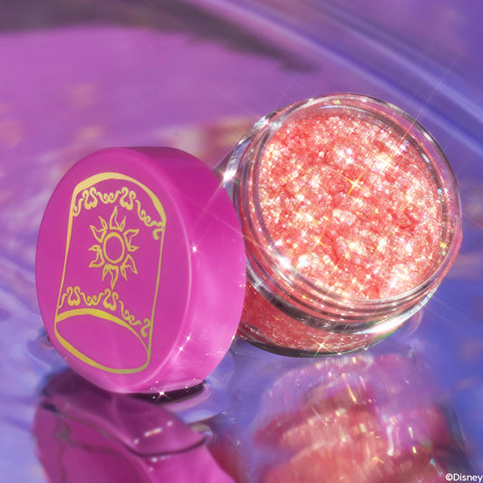 Open jar of ColourPop Glitter Gel, Floating Lights, gleams with peachy-pink iridescent flecks; the clear pot rests on a glossy lavender reflective surface with a magenta lid featuring a gold sun motif beside it.